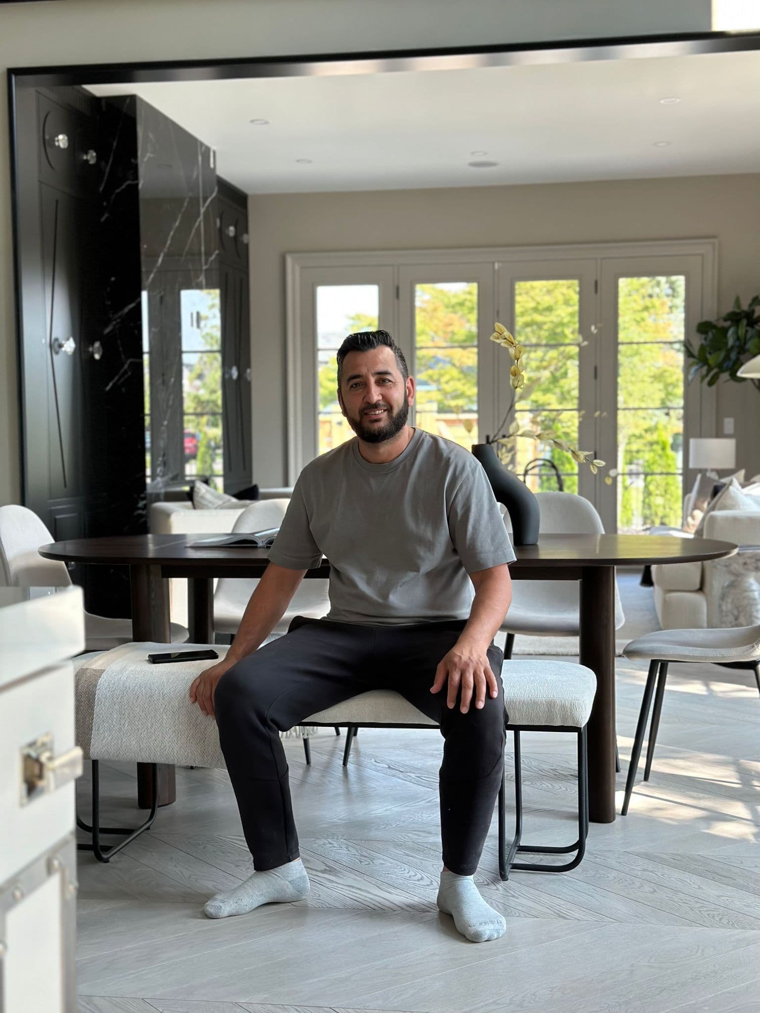 Smiling bearded man sits on a white bench in a bright, modern dining room.