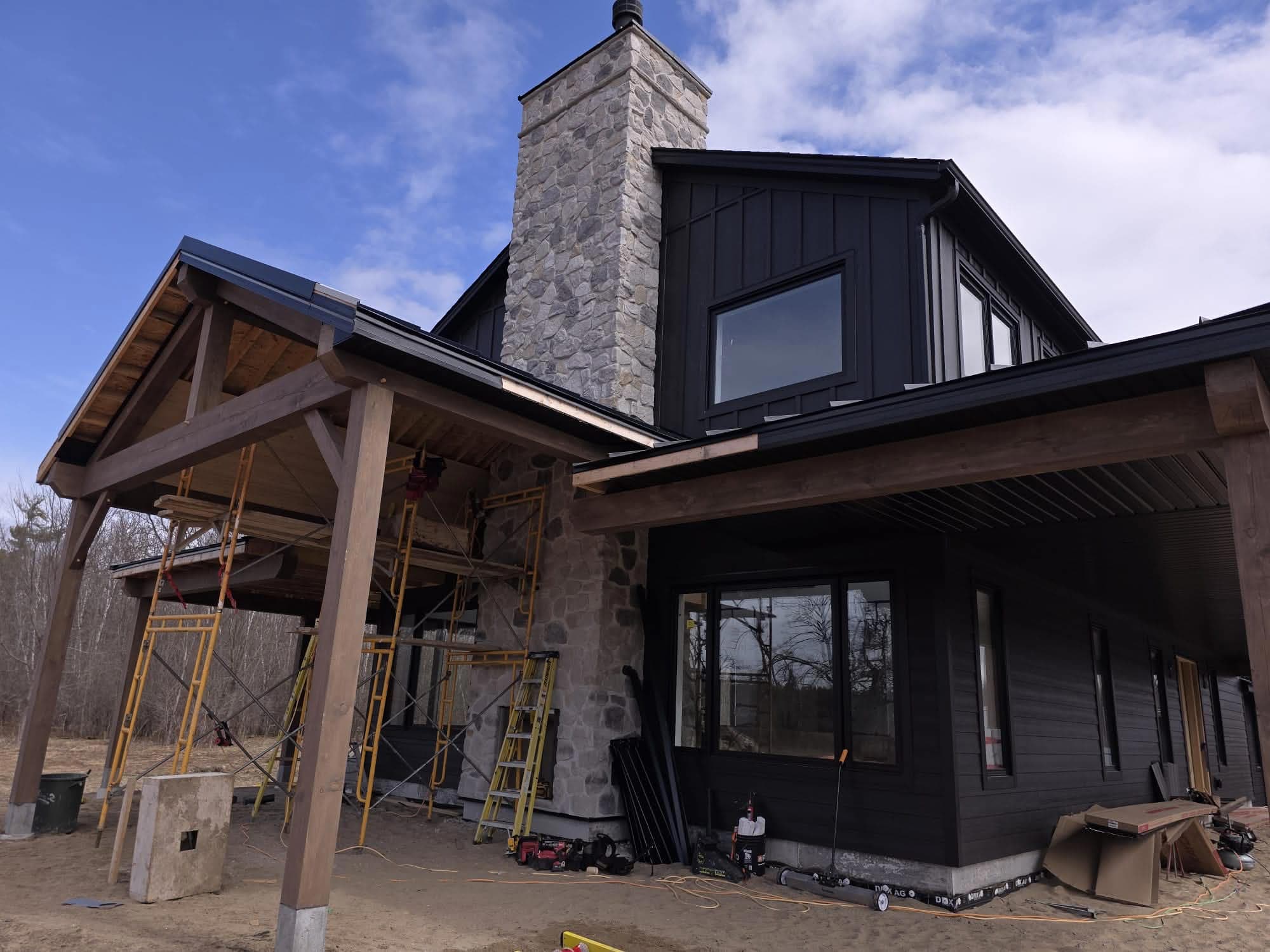 Modern black house under construction with a tall stone chimney and timber-framed porch structure.